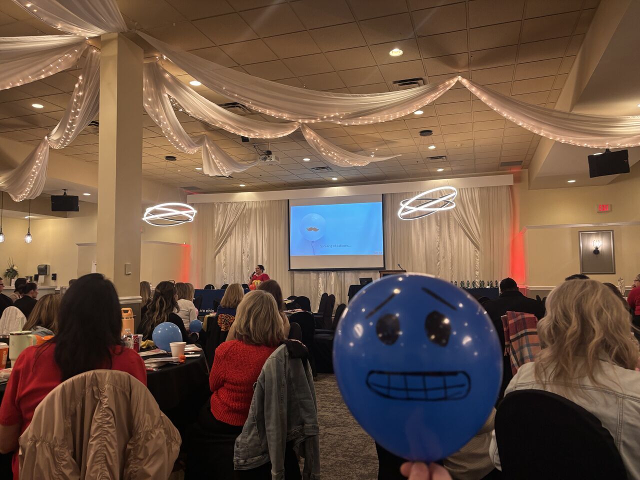 Amy C. Waninger presents from the stage in the background. In the midground, audience members watch intently from their tables. In the foreground, an audience member holds up a blue ballon with a worried face drawn on it. This balloon represents the audience member's feeling about change.